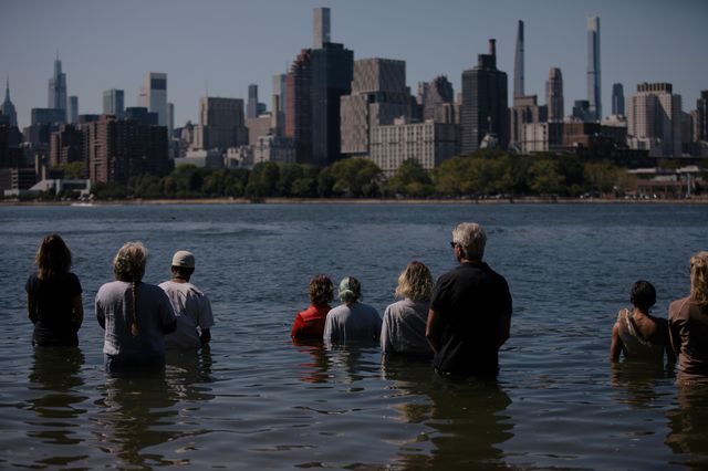 Performance artist Sarah Cameron Sunde (red) is joined by residents and supporters in the fourth hour of a performance piece where she will stand on the shores the East River at Hallet’s Cove for a full tidal cycle.